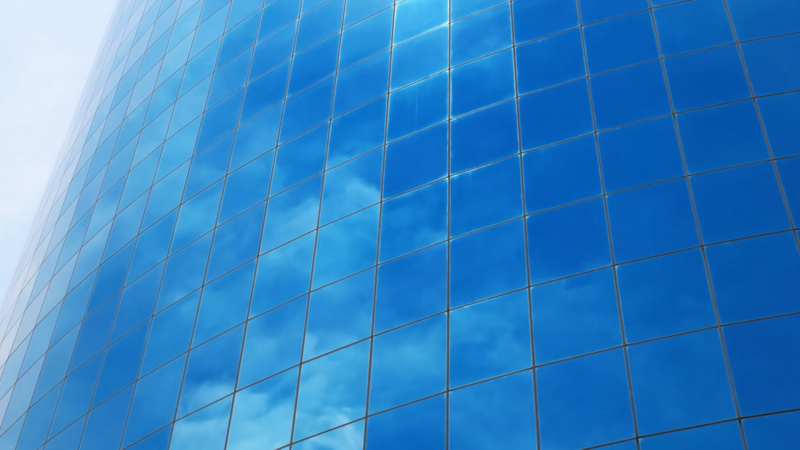 Modern glass office building with reflective blue windows mirroring a cloudy sky, viewed from a low angle to emphasize the buildings height and geometric grid pattern.
